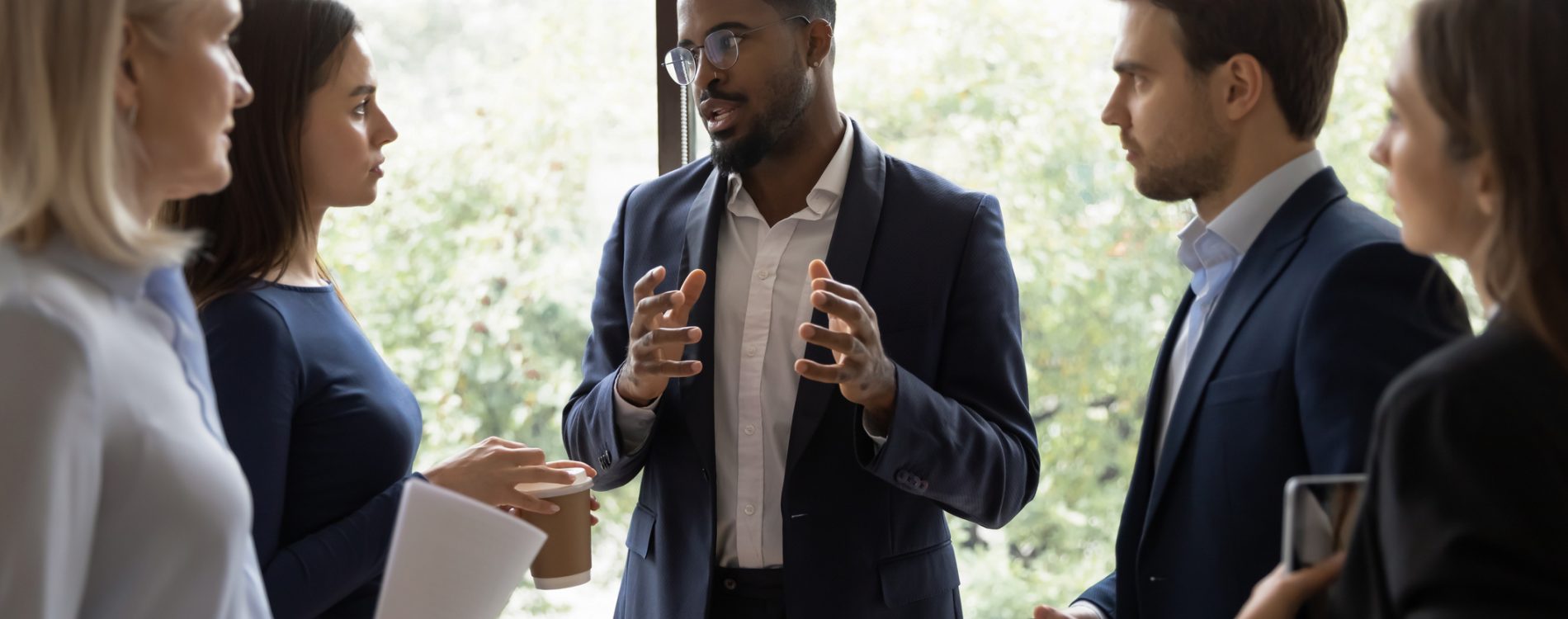 Confident African male leader standing telling diverse colleagues about new project, boss lead briefing for different age workgroup team employees in office. Sharing information and leadership concept